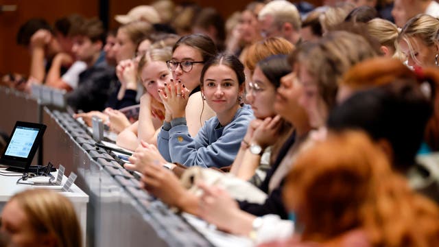 Eine Gruppe von Studierenden sitzt in einem Hörsaal. Eine junge Frau in der Mitte mit einem blauen Pullover schaut direkt in die Kamera. Vor ihr befindet sich ein Tablet mit Notizen. Die Atmosphäre ist aufgekratzt und lernorientiert.