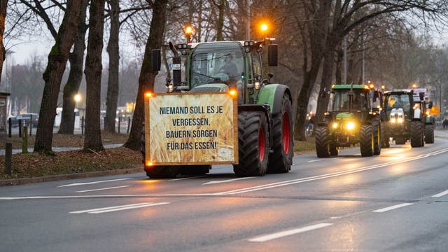 Mehrere Traktoren fahren hintereinander auf der Straße. Der vorderste hat ein Schild an der Stoßstange, auf dem steht: Niemand soll es je vergessen, Bauern sorgen für das Essen.