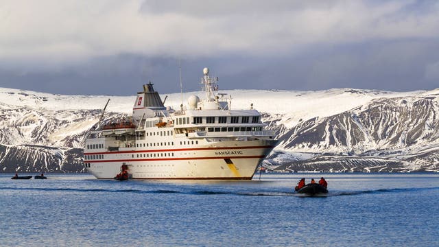 Ein großes Kreuzfahrtschiff mit dem Namen "Hanseatic" fährt durch eine ruhige, eisige Gewässerlandschaft, umgeben von schneebedeckten Bergen. Im Vordergrund bewegt sich ein kleines Schlauchboot mit mehreren Personen in Richtung des Schiffes. Der Himmel ist bewölkt, was der Szene eine kühle, arktische Atmosphäre verleiht.