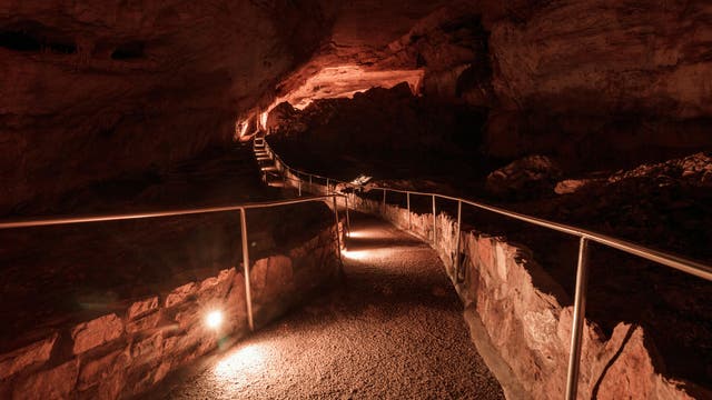 Ein beleuchteter Weg führt durch die Carlsbad Caverns in New Mexico. Der Weg ist grobkörnig und grau und von gemauerten Steinen eingefasst, ein Handlauf aus Stahl mit stählernen Pfosten führt an ihm entlang. Die Höhlendecke wirkt im Licht rötlich. Insgesamt ist es düster.