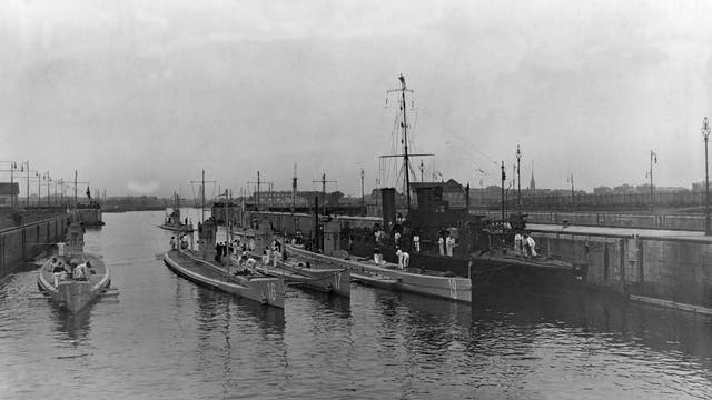 Historische Schwarz-Weiß-Fotografie eines Hafens mit mehreren U-Booten und einem Schiff, die an einem Kai festgemacht sind. Auf den U-Booten und dem Schiff sind mehrere Personen in Uniformen zu sehen, die verschiedene Aufgaben ausführen. Im Hintergrund sind industrielle Gebäude und Kräne zu erkennen, die auf eine geschäftige Hafenumgebung hindeuten. Die Szene vermittelt einen Eindruck von maritimer Aktivität und militärischer Präsenz.