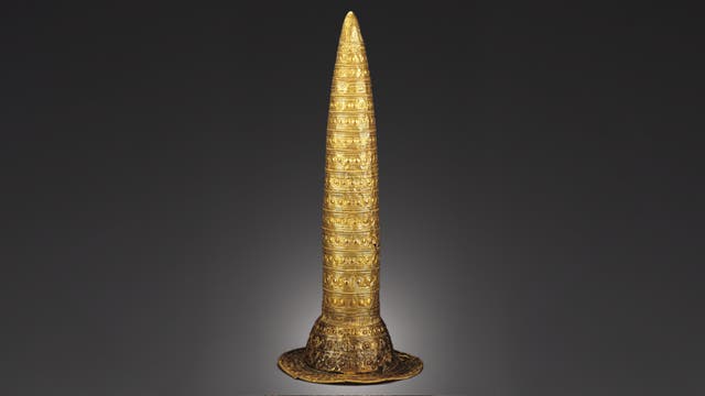 Ein konischer, goldener Hut mit detaillierten, kreisförmigen und linearen Mustern, die die Oberfläche bedecken. Der Hut hat eine breite Krempe und ist auf einem neutralen, dunklen Hintergrund abgebildet. Die Verzierungen sind symmetrisch und gleichmäßig verteilt, was auf eine kunstvolle Handwerkskunst hinweist.