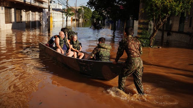 Menschen werden aufgrund der Überschwemmungen durch Mitarbeiter der brasilianischen Streitkräfte mit einem Boot evakuiert.  In den Straßen stehen braune Fluten, immerhin scheint gerade die Sonne.