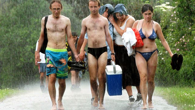 Eine Gruppe von sechs Personen geht in Badebekleidung durch starken Regen auf einem nassen Weg. Zwei Männer tragen eine Kühlbox und Schuhe in der Hand. Eine Frau hält ein Handtuch und bedeckt ihren Kopf mit einem Kleidungsstück. Die Szene vermittelt einen unerwarteten Regenschauer, der zum Abbruch eines Badeausflugs führt.