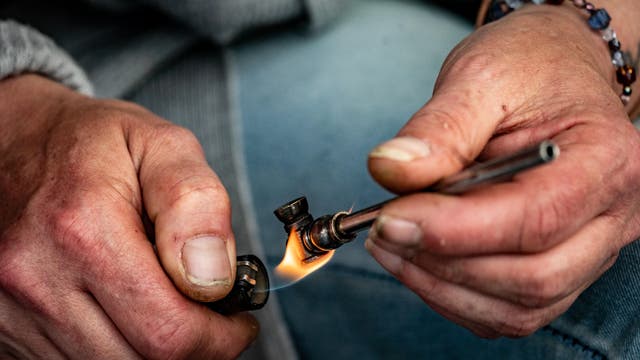 Eine Person hält eine kleine Metallpfeife in der rechten Hand und zündet sie mit einem Feuerzeug in der linken Hand an. Die Hände sind nah aufgenommen, und die Person trägt ein Armband mit bunten Perlen. Die Szene vermittelt den Akt des Rauchens.