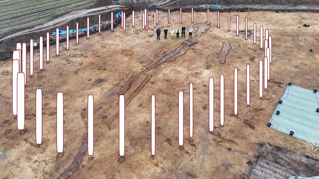 Eine archäologische Ausgrabungsstätte mit einem großen, kreisförmigen Bereich, der durch weiße Markierungen im Bild hervorgehoben ist. In der Mitte stehen mehrere Personen, die die Ausgrabung inspizieren. Der Boden zeigt Spuren von Pfostenlöchern. Im Hintergrund sind landwirtschaftliche Felder zu sehen. Die Szene vermittelt den Eindruck einer bedeutenden archäologischen Entdeckung.