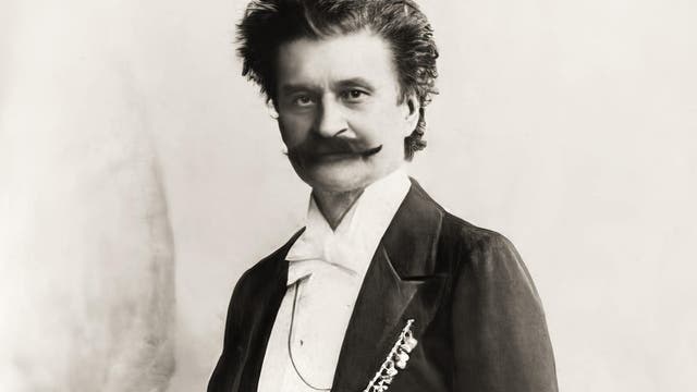 Ein schwarz-weißes Porträtfoto eines Mannes in formeller Kleidung mit durchdringendem Blick. Er trägt einen Anzug mit Fliege und hat einen markanten Schnurrbart. Der Hintergrund ist neutral und schlicht, was den Fokus auf die Person lenkt.