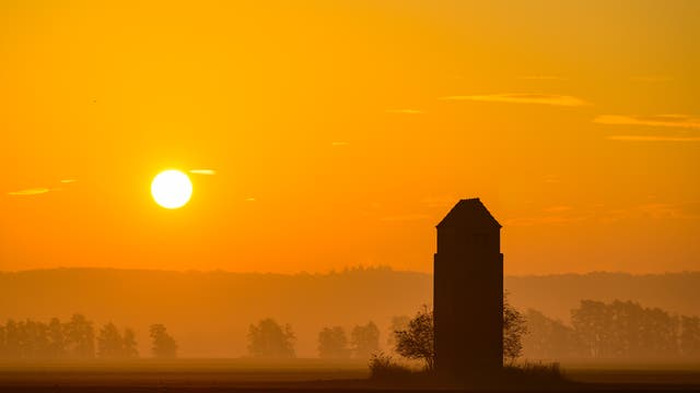 Die Sonne geht als gleißende helle Scheibe im oderbruch auf, der Himmel ist orange. Im Vordergund ist dunkel ein Turm erkennbar. Im Hintergrund befinden sich Bäume im Morgendunst, ganz hinten zeichnet sich ein Höhenzug ab