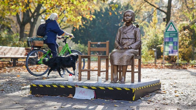 Eine Bronzestatue eines sitzenden Mädchens auf einem Podest in einem herbstlichen Park. Neben der Statue steht ein leerer Stuhl. Im Hintergrund fährt eine Person auf einem grünen Fahrrad vorbei, begleitet von einem schwarzen Hund. Auf dem Podest ist ein handgeschriebenes Schild mit der Aufschrift "ARI bleibt!" angebracht. Bäume mit herbstlichem Laub und ein Informationsschild sind im Hintergrund zu sehen.