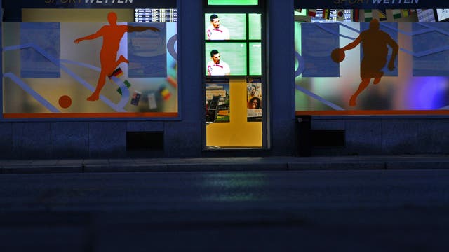 Das Foto zeigt ein erleuchtetes Wettbüro in einer nächtlichen Straße. Durch die offene oder gläserne Tür sieht man Monitore mit einer Sportübertragung.