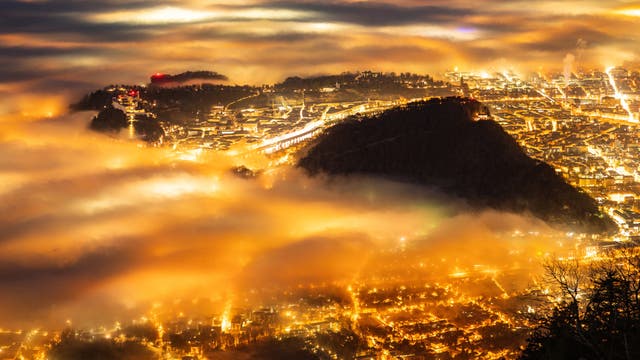 Eine nächtliche Stadtansicht, die von oben aufgenommen wurde, zeigt eine Stadt, die in warmes, goldenes Licht getaucht ist. Dichter Nebel schwebt über der Stadt und erzeugt eine mystische Atmosphäre. Die Lichter der Stadt leuchten durch den Nebel hindurch, während im Hintergrund Hügel oder Berge zu sehen sind. Die Szene vermittelt ein Gefühl von Ruhe und Erhabenheit.