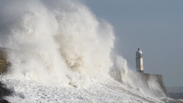 Hohe Wellen schlagen gegen eine Küstenmauer, neben der ein weißer Leuchtturm steht. Die Gischt spritzt dramatisch in die Luft, was die Kraft des Meeres verdeutlicht. Der Himmel ist grau und wolkenverhangen, was auf stürmisches Wetter hindeutet.