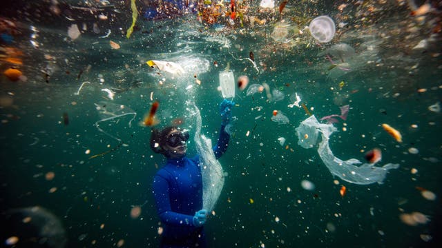 Eine Person in einem blauen Taucheranzug und einer Tauchmaske schwimmt unter Wasser inmitten von Plastikmüll. Sie hält ein Stück Plastik in der Hand, umgeben von verschiedenen Abfällen wie Plastiktüten und Flaschen. Das Bild thematisiert die Verschmutzung der Ozeane durch Plastikmüll.