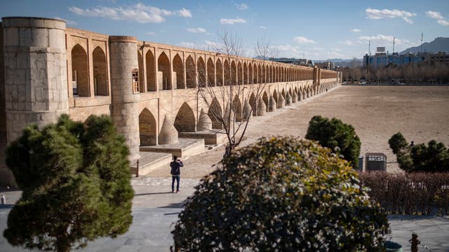 Eine historische Brücke mit mehreren Bögen erstreckt sich über ein trockenes Flussbett. Die Brücke besteht aus hellem Stein und zeigt eine Reihe von Arkaden. Im Vordergrund sind Bäume und ein Mensch, der die Brücke fotografiert. Der Himmel ist klar mit einigen Wolken, und im Hintergrund sind Gebäude und Berge zu sehen.