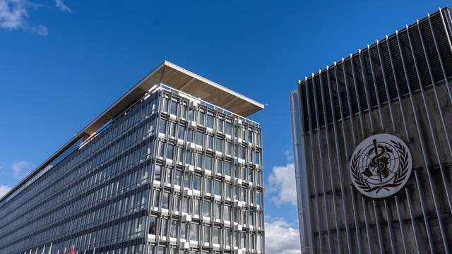 Zwei moderne Bürogebäude unter einem klaren blauen Himmel. Links steht ein hohes Gebäude mit vielen Fenstern und einer markanten Dachkante, rechts ein Gebäude mit dem Logo der Weltgesundheitsorganisation (WHO) auf der Fassade. Vor dem linken Gebäude sind mehrere internationale Flaggen zu sehen.