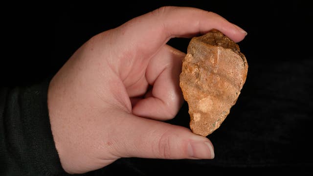 Eine Hand hält ein steinernes Artefakt, das wie eine Pfeilspitze geformt ist, vor einem schwarzen Hintergrund. Auf der Oberfläche des Artefakts sind natürliche Texturen und Farbvariationen zu erkennen. 