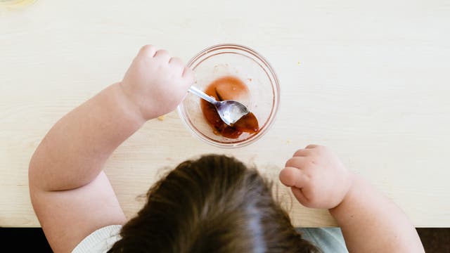 Blick von oben auf den Kopf eines Kindes mit braunen Haaren, das an an einem Holztisch sitzt und mit einem Löffel aus einer kleinen Glasschüssel eine bräunliche, an Götterspeise erinnernde Nachspeise isst. Auf dem Tisch im oberen linken Eck steht ein Glas mit einem gelben Getränk.