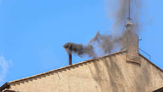 Ein Schornstein auf dem Dach der Sixtinischen Kapelle stößt schwarzen Rauch in den blauen Himmel aus.