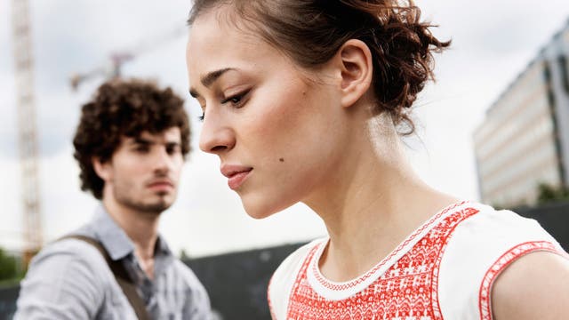 Eine junge Frau im Vordergrund blickt bedrückt nach unten, während ein Mann im Hintergrund, der sie anschaut, nur unscharf zu sehen ist. Die Frau trägt ein weißes Oberteil mit rotem Muster. Im Hintergrund sind Baukräne und ein modernes Gebäude sichtbar, was auf eine städtische Umgebung hindeutet. Die Szene vermittelt eine nachdenkliche bis krisenhafte Stimmung.