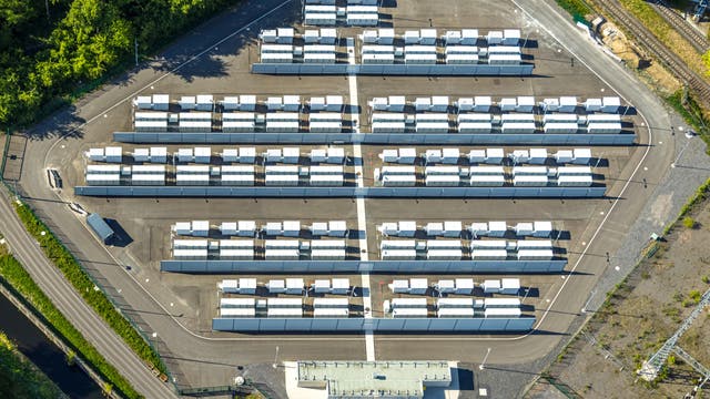 Luftaufnahme eines großen Solarkraftwerks mit mehreren Reihen von Solarmodulen, die auf einem asphaltierten Gelände angeordnet sind. Die Module sind in parallelen Linien aufgestellt, umgeben von Straßen und Vegetation. Im Vordergrund befindet sich ein Gebäude, das möglicherweise als Kontrollzentrum dient. Die Szene zeigt eine klare, strukturierte Anordnung der Solarpanels, die auf nachhaltige Energieerzeugung hinweist.