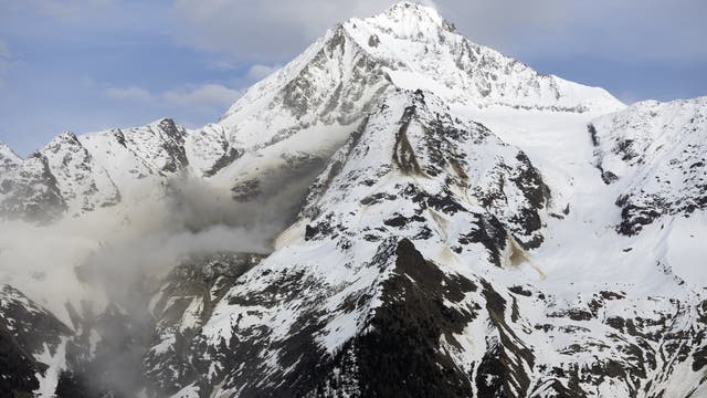 Schneebedeckter Berggipfel unter blauem Himmel mit einigen Wolken. Der Berg ist von steilen, schneebedeckten Hängen umgeben, die teilweise von dunklen Felsen durchzogen sind. Eine leichte Nebelbildung ist in der linken Bildhälfte zu sehen. Die Szene vermittelt eine ruhige, majestätische Atmosphäre in einer alpinen Landschaft.