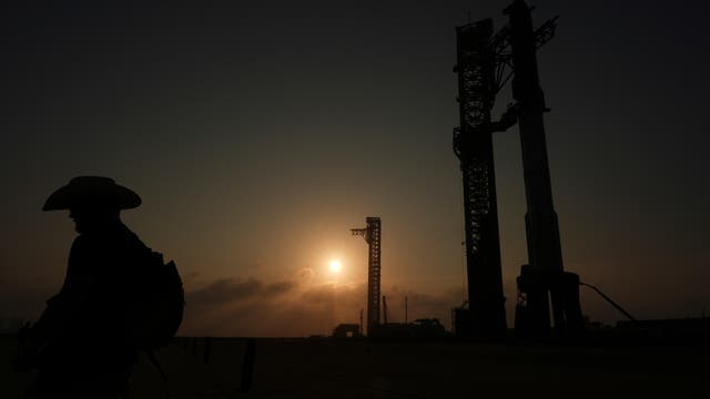 Ein Sonnenuntergang mit der Silhouette eines Mannes mit Cowboyhut und Rucksack. Rechts im Bild sind zwei große, turmartige Strukturen einer Raketenabschussrampe zu sehen. 