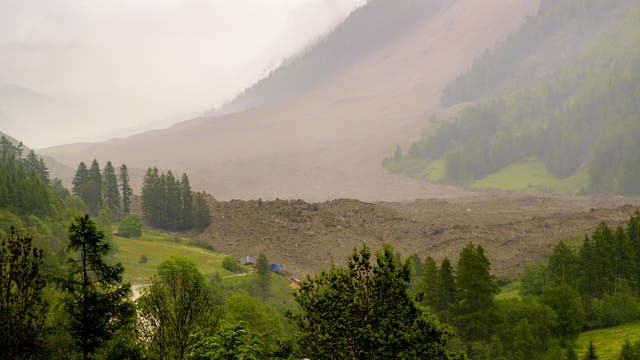Eine weitläufige Landschaft mit einem großen Erdrutsch, der einen Hang hinunterfließt. Im Vordergrund sind grüne Bäume und Wiesen zu sehen, während der Himmel bewölkt ist und Nebel die Berge umhüllt. Keine Menschen und nur wenige Gebäude sind sichtbar.