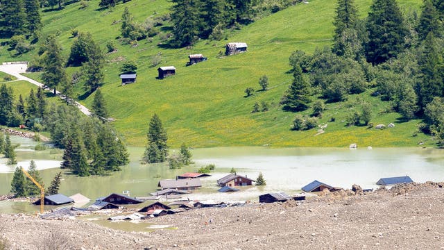 Eine ländliche Landschaft in den Schweizer Alpen mit grünen Hügeln und vereinzelten Bäumen. Im Vordergrund sind mehrere Gebäude teilweise von Hochwasser überflutet. Die Dächer der Häuser ragen aus dem Wasser hervor. Im Hintergrund sind kleine Holzhütten auf einer grünen Wiese zu sehen. Ein Kran ist links im Bild sichtbar. 