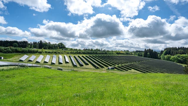 Eine weitläufige Solaranlage auf einem grünen Hügel unter einem bewölkten Himmel. Die Solarpaneele sind in geordneten Reihen angeordnet und erstrecken sich über die Landschaft. Im Hintergrund sind Bäume und ein paar Gebäude zu sehen. Die Szene vermittelt ein Gefühl von nachhaltiger Energieerzeugung und Umweltschutz.