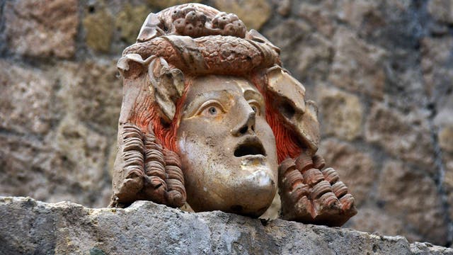 Steinerne Skulptur eines Kopfes mit ausdrucksstarkem Gesicht, roten Haaren und einem kunstvoll verzierten Kopfschmuck, der Früchte darstellt. Die Skulptur ist auf einer Steinmauer montiert, die als Hintergrund dient.
