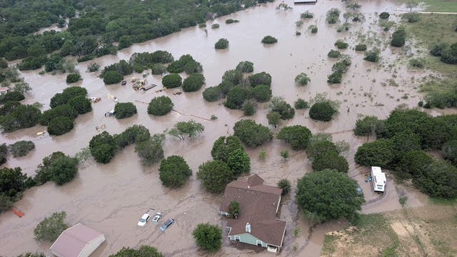 Luftaufnahme einer überfluteten Landschaft in Texas am Guadalupe River mit mehreren Häusern und Fahrzeugen, die teilweise im Wasser stehen. Bäume ragen aus dem Wasser heraus, während die Umgebung von braunem Hochwasser bedeckt ist. Im Vordergrund ist ein Haus mit braunem Dach zu sehen, daneben ein Wohnwagen. 