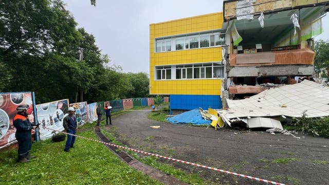 Ein teilweise eingestürztes Gebäude mit gelber und blauer Fassade. Der rechte Teil des Gebäudes ist stark beschädigt, mit herabgefallenen Trümmern und freiliegenden Innenräumen. Im Vordergrund stehen mehrere Personen in Schutzkleidung hinter einer Absperrung. Links sind Bäume und ein Zaun mit Plakaten zu sehen. Der Himmel ist bewölkt.