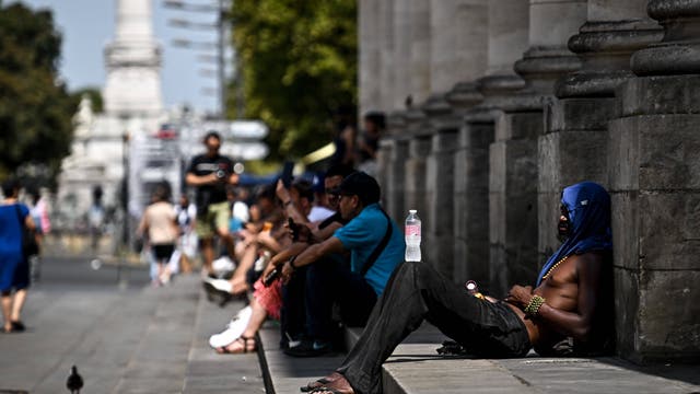 Eine Gruppe von Menschen sitzt auf den Stufen eines großen Gebäudes im Freien. Eine Person im Vordergrund lehnt sich mit einer Wasserflasche auf dem Bein zurück. Die Szene zeigt einen sonnigen, heißen Tag in einer städtischen Umgebung mit Bäumen und einer Statue im Hintergrund. Die Menschen scheinen sich auszuruhen oder zu erholen.
