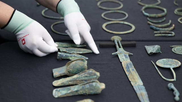 Eine Person mit weißen Handschuhen zeigt auf eine Sammlung alter Bronzeobjekte, die auf einem dunklen Tuch ausgebreitet sind. Zu den Artefakten gehören ein Schwert, Ringe und verschiedene Werkzeuge. Die Szene deutet auf eine archäologische Untersuchung oder Präsentation hin.