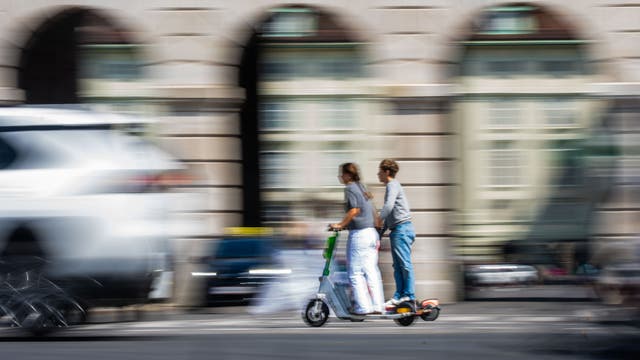 Zwei Personen fahren gemeinsam auf einem E-Scooter durch eine belebte Stadtstraße. Die Umgebung ist verschwommen, was die Bewegung und Geschwindigkeit betont. Im Hintergrund sind unscharfe Gebäude und vorbeifahrende Autos zu sehen.
