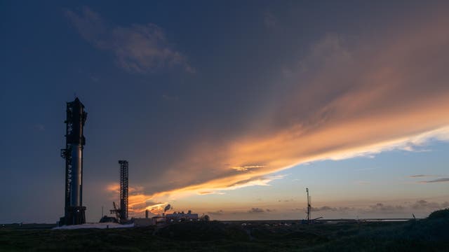 Eine Rakete steht auf einer Startrampe bei Sonnenuntergang. Der Himmel ist in warmen Orange- und Blautönen gefärbt, während Wolken den Horizont durchziehen. Die Szene vermittelt eine ruhige und erwartungsvolle Atmosphäre. Im Vordergrund ist die Silhouette der Rakete deutlich sichtbar, umgeben von technischen Strukturen.