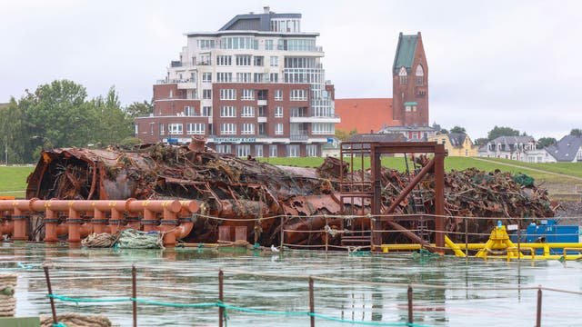 Ein altes, rostiges Schiffswrack liegt im Wasser vor einer modernen Wohnanlage und einem historischen Kirchturm. Im Vordergrund sind industrielle Rohre und Strukturen zu sehen. Die Szene zeigt einen Kontrast zwischen der verfallenen Struktur und der urbanen Umgebung im Hintergrund.