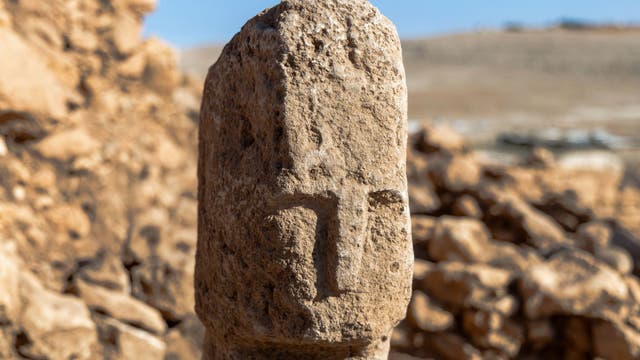 Ein alter, verwitterter Steinpfeiler mit eingraviertem, stark abstrahiertem Gesicht steht im Vordergrund einer archäologischen Ausgrabungsstätte. Der Hintergrund zeigt blauen Himmel und Landschaft. Der Fokus liegt auf den Details der Steinstruktur und der Gravur.