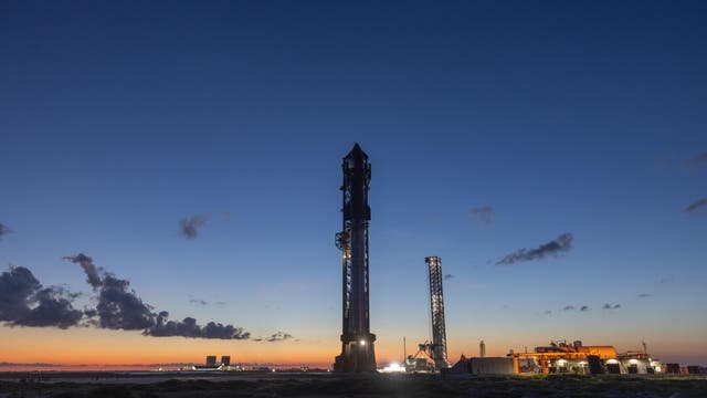 Eine Rakete steht auf einer Startrampe bei Sonnenuntergang. Der Himmel ist in tiefem Blau und Orange gefärbt. Im Vordergrund sind technische Anlagen und Strukturen zu sehen, die beleuchtet sind. Wolken ziehen am Himmel vorbei, während die Szene eine ruhige, aber erwartungsvolle Atmosphäre vermittelt.