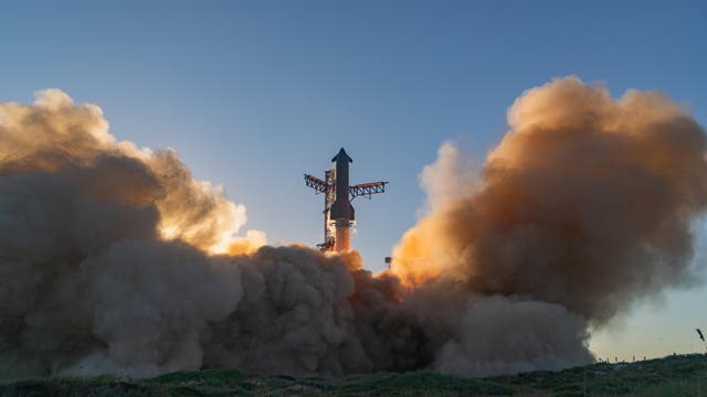 Eine Rakete hebt von einer Startrampe ab, umgeben von dichten Rauch- und Staubwolken. Der Himmel ist klar und blau, während die Sonne im Hintergrund die Szene beleuchtet. Die Rakete ist vertikal ausgerichtet und von metallischen Strukturen umgeben.