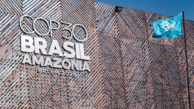Gebäude mit einer modernen, geometrischen Fassade, auf der in großen Buchstaben "COP30 BRASIL AMAZÔNIA BELÉM 2025" steht. Rechts weht eine blaue Flagge mit dem Logo der Vereinten Nationen. Der Himmel ist klar und blau.