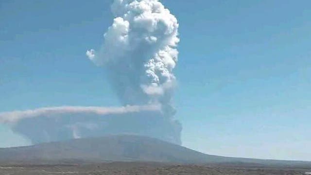 Ein Vulkan in einer kargen Landschaft stößt eine große, weiße Rauch- und Aschewolke in den klaren blauen Himmel aus. Die Wolke steigt hoch auf und breitet sich nach oben hin aus. Im Vordergrund ist eine flache, trockene Ebene zu sehen.