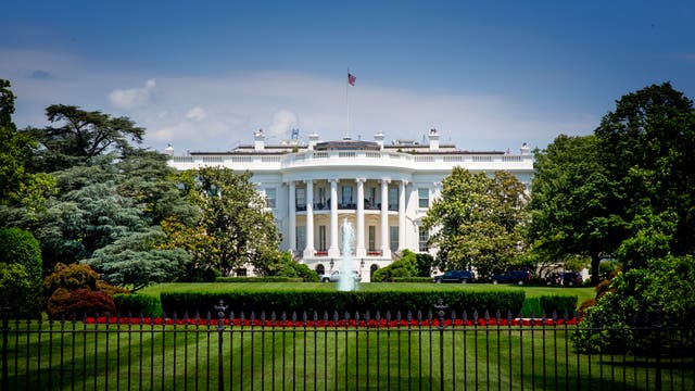 Das Bild zeigt das Weiße Haus in Washington, D.C., ein ikonisches Gebäude mit einer weißen Fassade und Säulen, umgeben von gepflegten Rasenflächen und Bäumen. Eine amerikanische Flagge weht auf dem Dach. Der Himmel ist blau mit einigen Wolken.