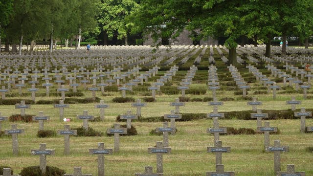 Ein weitläufiger Soldatenfriedhof mit zahlreichen grauen Steinkreuzen, die in geordneten Reihen aufgestellt sind. Im Hintergrund sind grüne Bäume zu sehen. Eine einzelne Person in blauer Kleidung geht zwischen den Gräbern entlang. Die Szene vermittelt eine ruhige und nachdenkliche Atmosphäre.