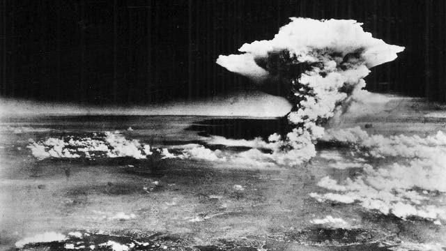 Eine Schwarz-Weiß-Fotografie zeigt eine große Pilzwolke, die in den Himmel aufsteigt, mit einer darunter liegenden Landschaft. Die Wolke besteht aus dichtem Rauch und Dampf, der sich nach oben ausbreitet. Die Szene vermittelt den Eindruck einer massiven Explosion. Im Vordergrund sind Teile einer Stadt und Küstenlinie zu erkennen.