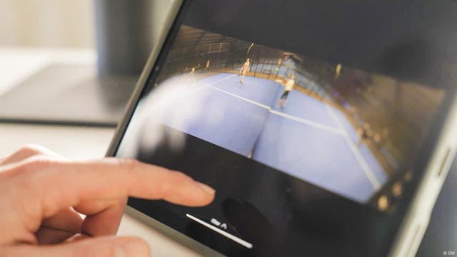 Eine Person bedient ein Tablet, auf dem ein Video von einem Tennisspiel auf einem blauen Platz abgespielt wird. Im Hintergrund sind zwei Spieler zu sehen. Die Hand der Person berührt den Bildschirm, um das Video zu steuern. Das Bild vermittelt den Eindruck von Technologie und Sport.