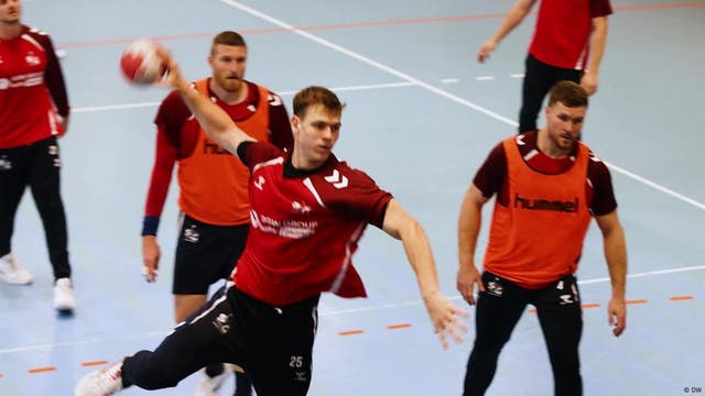 Eine Gruppe von Handballspielern in roten Trikots trainiert in einer Sporthalle. Ein Spieler im Vordergrund ist in der Bewegung des Wurfs, während andere Spieler im Hintergrund stehen und zuschauen. Der Boden ist hellblau mit markierten Linien.