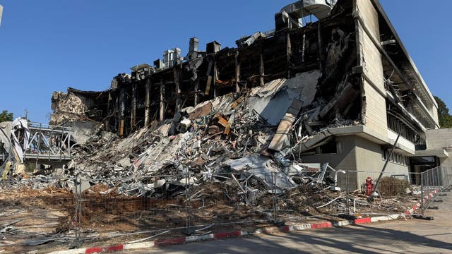 Ein teilweise eingestürztes Gebäude mit sichtbaren Trümmern und Stahlträgern. Die Fassade ist stark beschädigt, und es sind zahlreiche Schuttteile zu sehen. Ein Bauzaun umgibt die Ruine, und im Vordergrund ist eine Straße mit einem Zebrastreifen erkennbar. Der Himmel ist klar und blau.