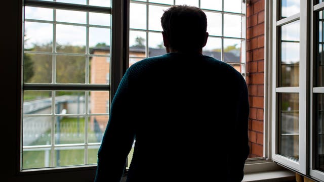 Eine Person steht mit dem Rücken zur Kamera und schaut aus einem großen, vergitterten Fenster in einen Hof mit Bäumen und einem Gebäude.