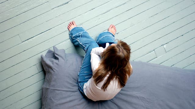 Eine Jugendliche mit langen, glatten, rotblonden Haaren sitzt auf einer grau bezogenen Matratze, die auf einem hellgrau gestrichenen Dielenboden liegt. Sie trägt eine Bluejeans und ein weißes Hemd und hat das Gesicht in den Händen vergraben. Die Perspektive ist so gewählt, dass man die triste Szene von oben, aus der Luft sieht. 